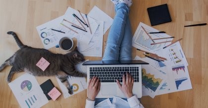 Ein junger Mitarbeiter einer Agentur sitzt auf dem Boden von Zetteln, Kaffee und einer Katze umgeben und arbeitet mit seinem Laptop auf dem Schoß.