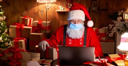 Weihnachtsmann mit Mund- und Nasenschutz vor einem Laptop, im Hintergrund ein Baum und Geschenke
