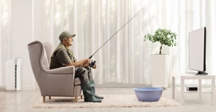 Ein Angler sitzt mit seiner Angelrute im Wohnzimmer vor einem Fernseher.