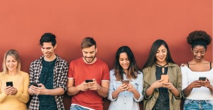 Sechs junge Menschen verschiedenen Geschlechts mit unterschiedlicher Hautfarbe stehen vor einer orangefarbenen Wand. Alle blicken auf ihr Smartphone und wollen Social Tokens kaufen.