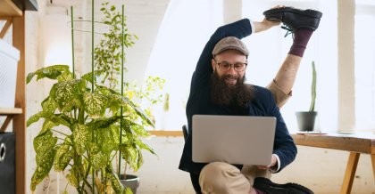 Ein junger Mann mit Bart sitzt mit dem Notebook auf einem Stuhl. Er dehnt sich und kann sein Bein hinter den Rücken biegen. Er ist ein flexibler Softwareentwickler für Unternehmen.