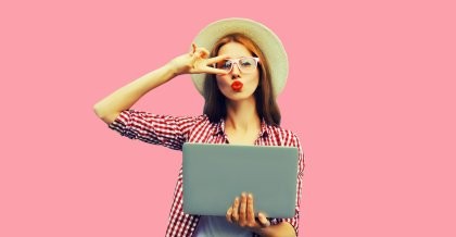 Junge Frau mit Hut und Brille vor rosarotem Hintergrund. Sie macht eine coole Geste. Ihr gefällt die UX Optimierung, die sie auf ihrem Notebook betrachtet.