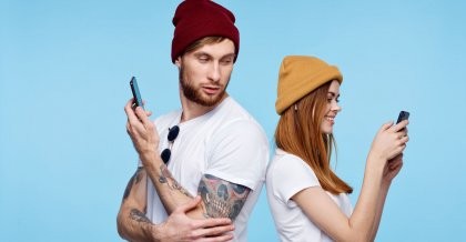 Ein junges Paar steht Rücken an Rücken, sie ist in ihr Smartphone vertieft, er schaut misstrauisch über die Schulter zu ihr. Automatisiert Technologie das Liebesleben?